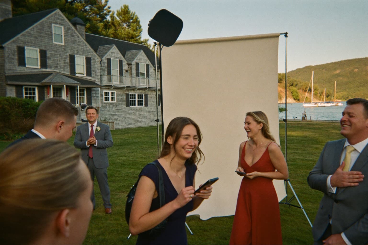 Wedding guests laughing at an ivory photo booth backdrop on a shoreline yacht-club lawn at golden hour.