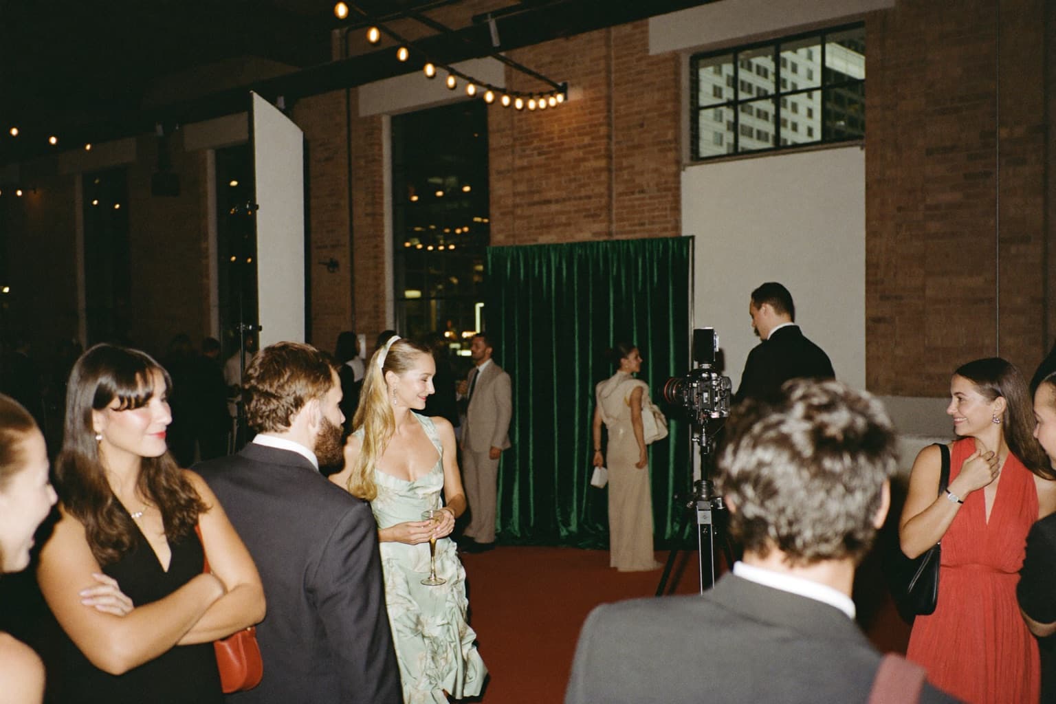 Guests mingling near an emerald velvet photo booth backdrop in an exposed-brick industrial event space at night.