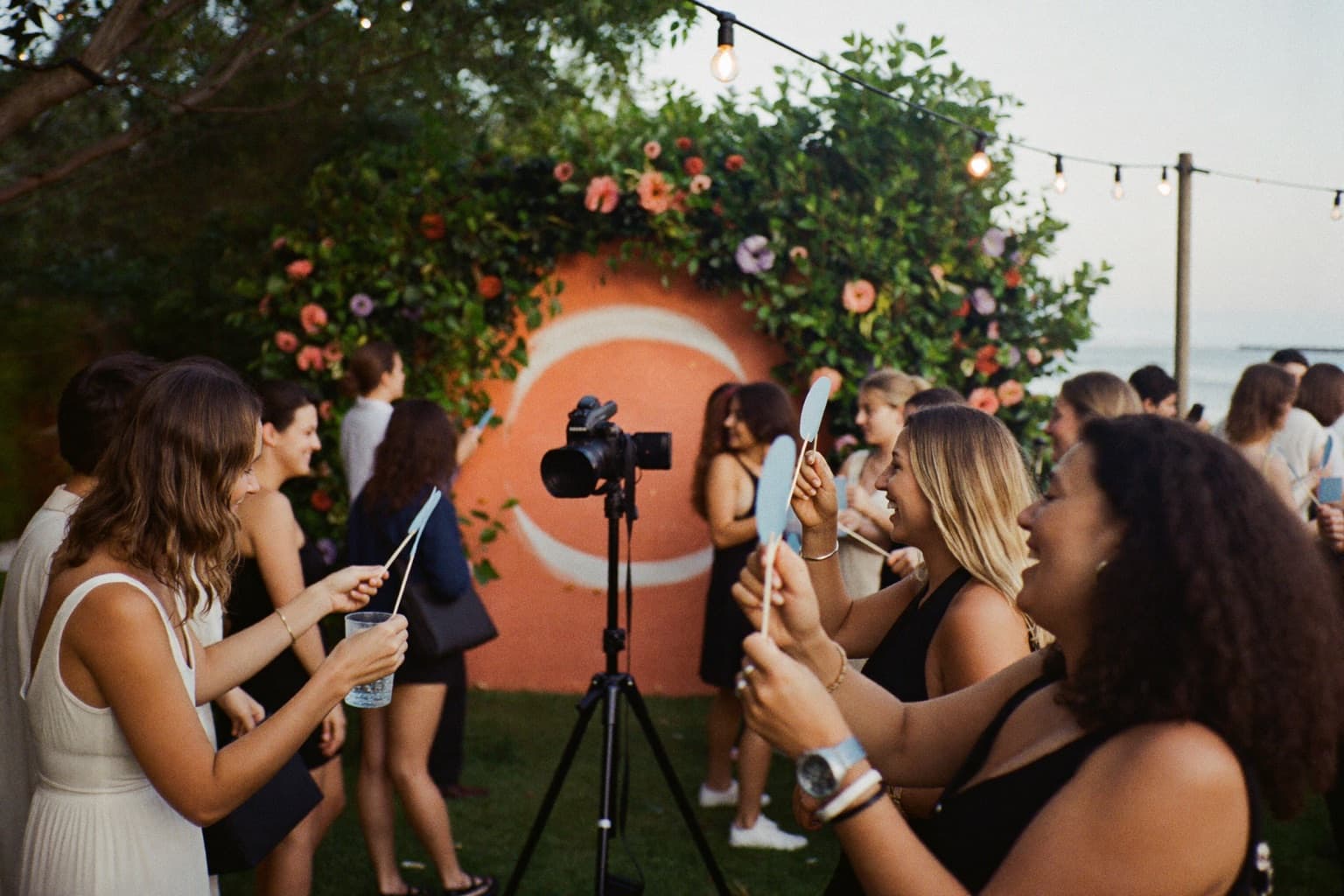 Guests holding props at an open-air photo booth with a floral backdrop during a coastal country-club celebration.