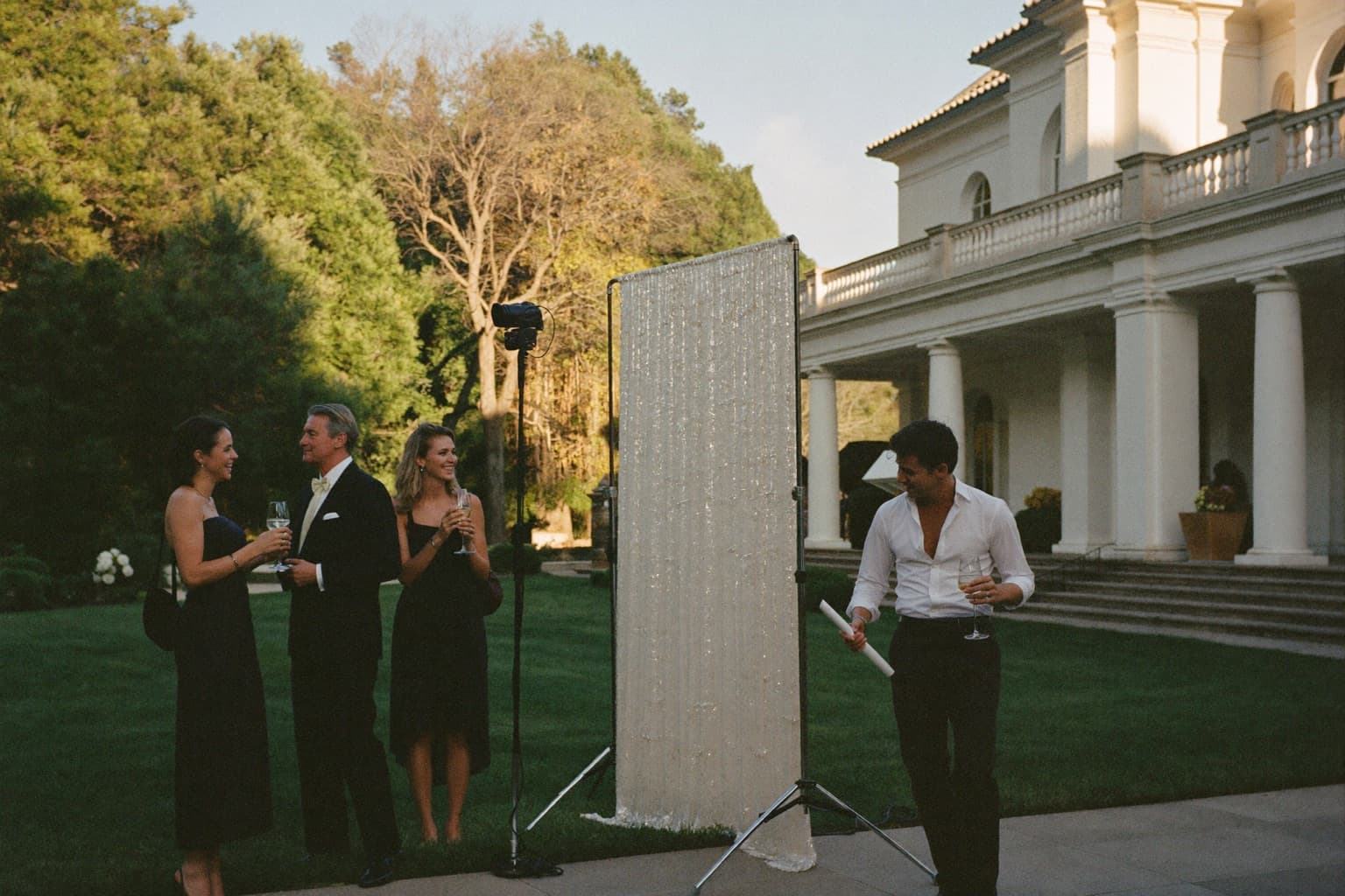 Guests with champagne at an ivory sequin photo booth backdrop on a Colonial Revival estate terrace.