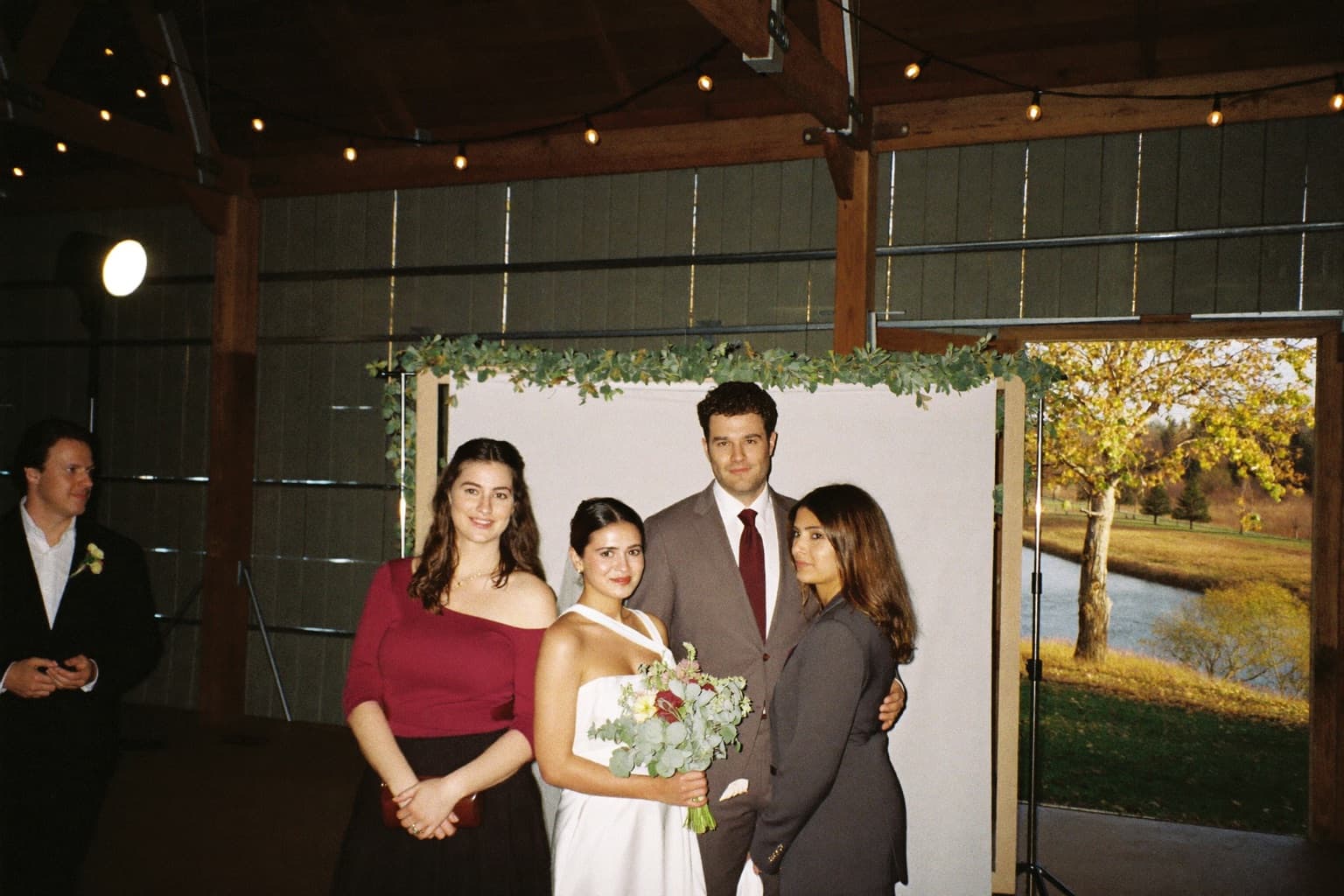 A wedding party at a garland-draped photo booth backdrop inside a post-and-beam barn in autumn.