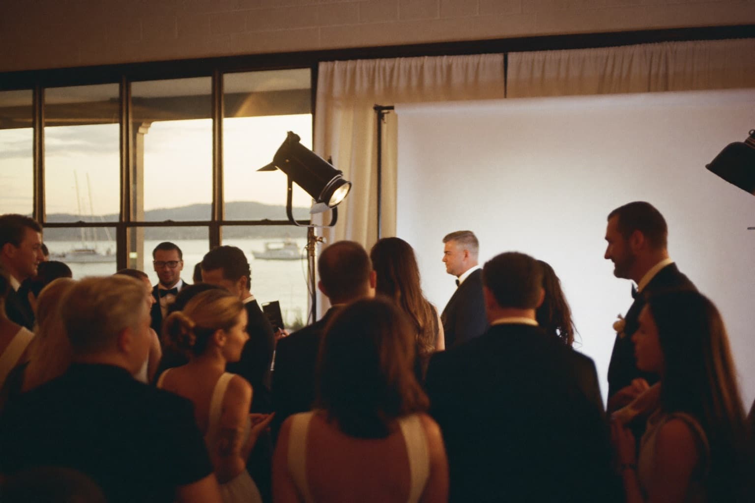 Guests in black-tie attire at a glam photo booth during a waterfront country-club wedding reception at golden hour.