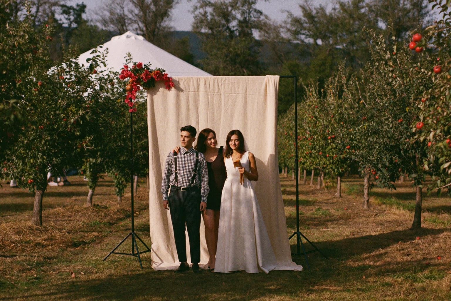Guests posing at a cream linen photo booth backdrop in an apple orchard in October light.