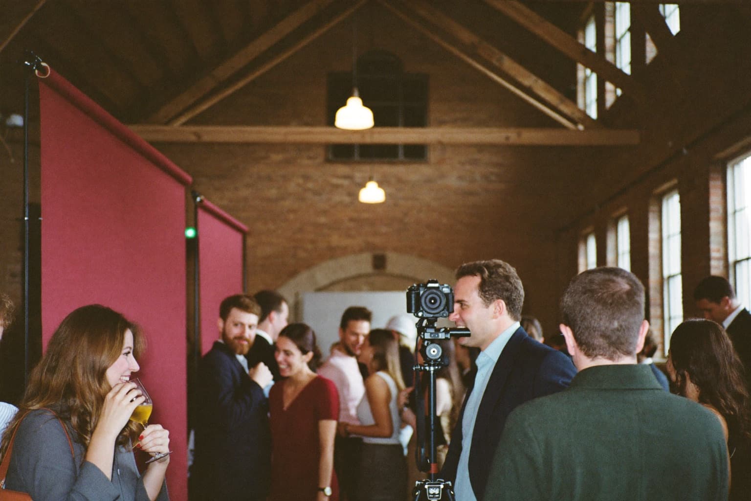 Guests at a deep-red photo booth backdrop inside a restored brick mill hall with timber trusses.