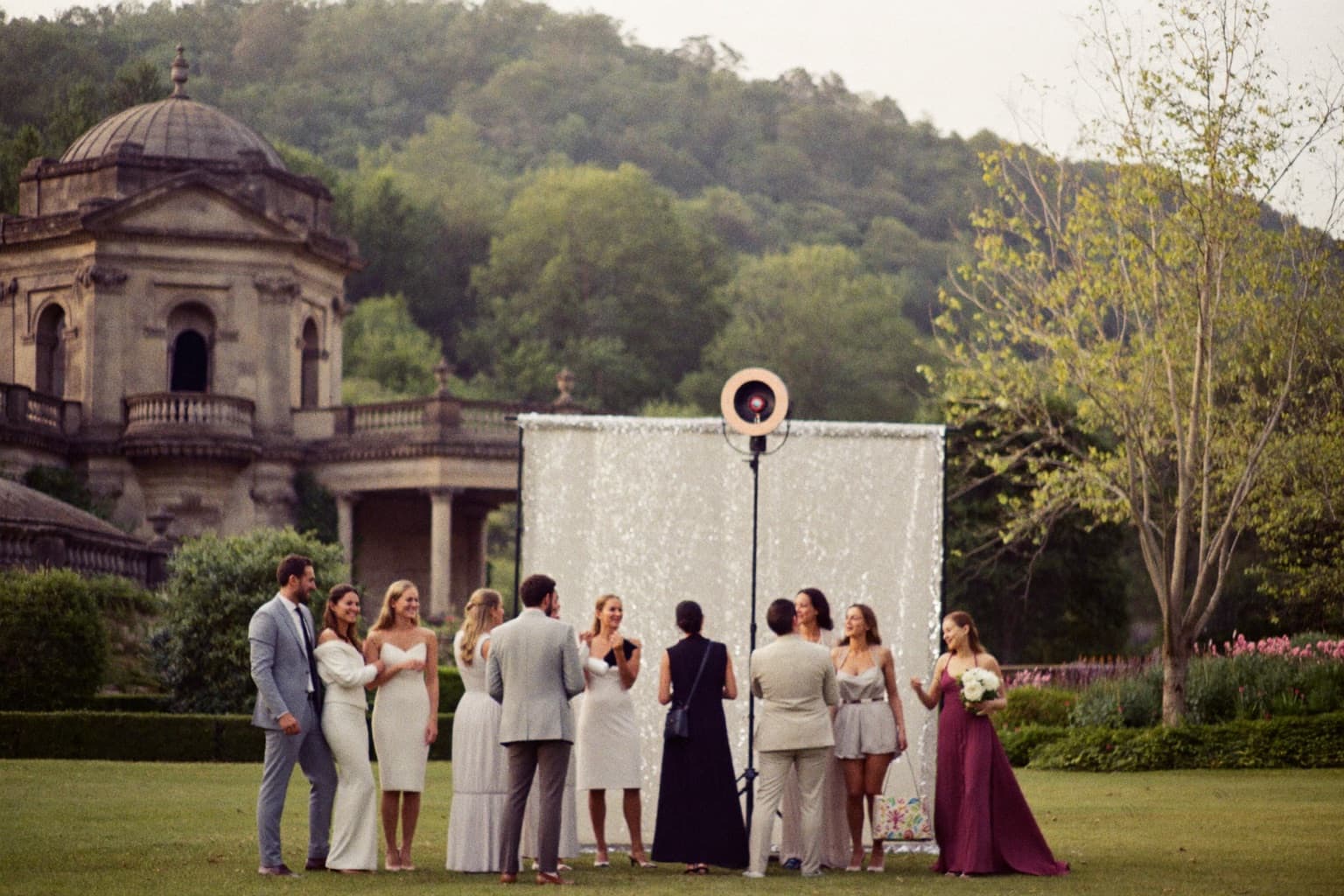 Guests gathered at a white sequin photo booth backdrop with a ring light beside a historic stone pavilion in a park.