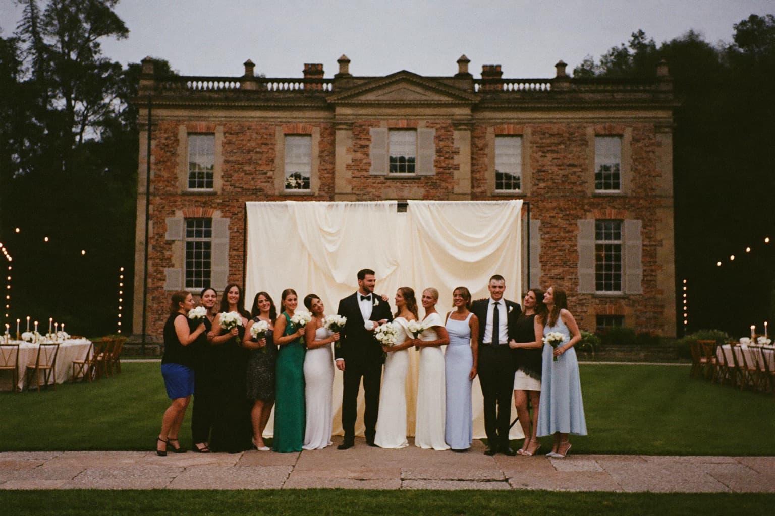 A wedding party posing at an ivory draped photo booth backdrop on the lawn of a grand historic brick mansion at dusk.