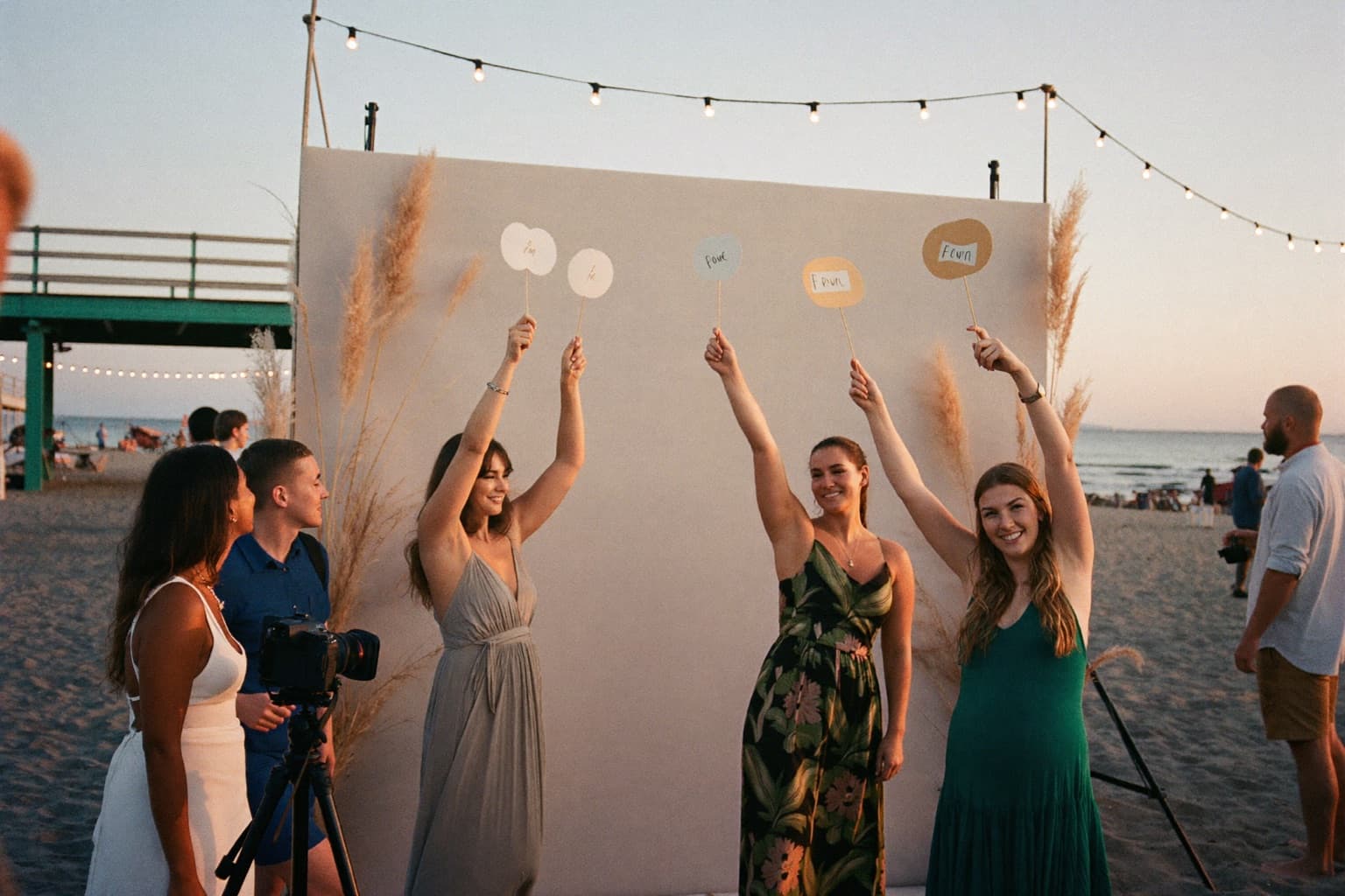 Guests holding props at an open-air photo booth backdrop under string lights at a shoreline pavilion at sunset.