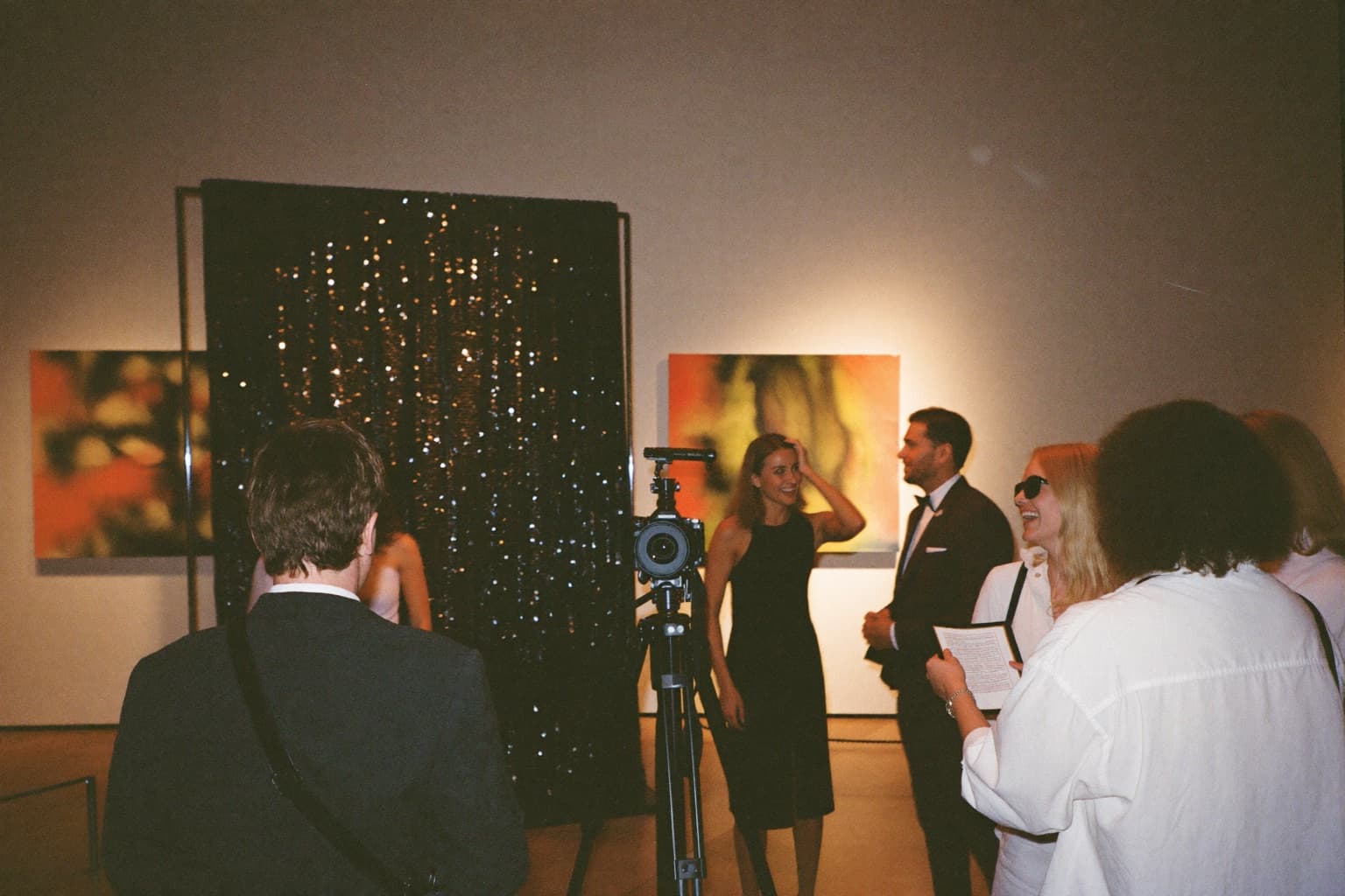 Guests in evening wear at a black sequin photo booth backdrop with a camera in a modern art gallery during a gala.