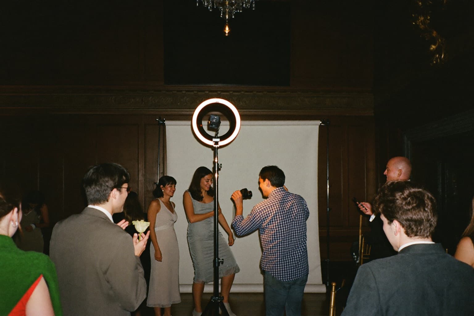 Guests laughing at an open-air photo booth with a white sequin backdrop in a historic ballroom during an evening wedding reception.