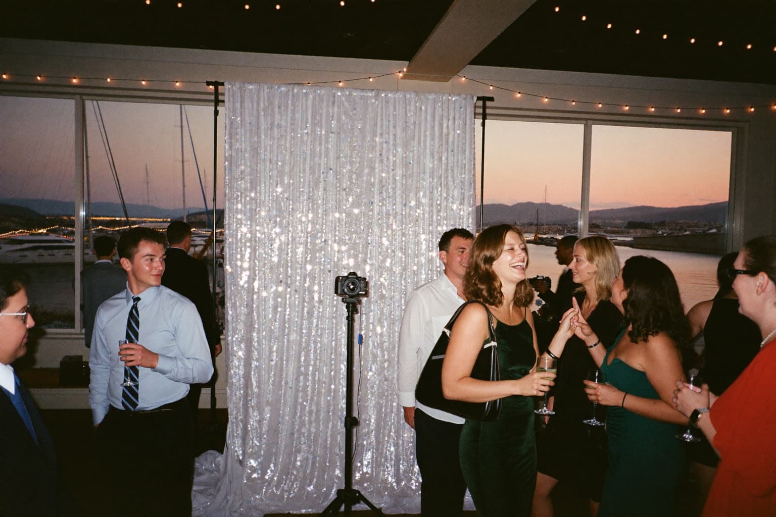 Guests laughing beside a silver sequin photo booth backdrop and camera in a harborside event room with sailboats at dusk.