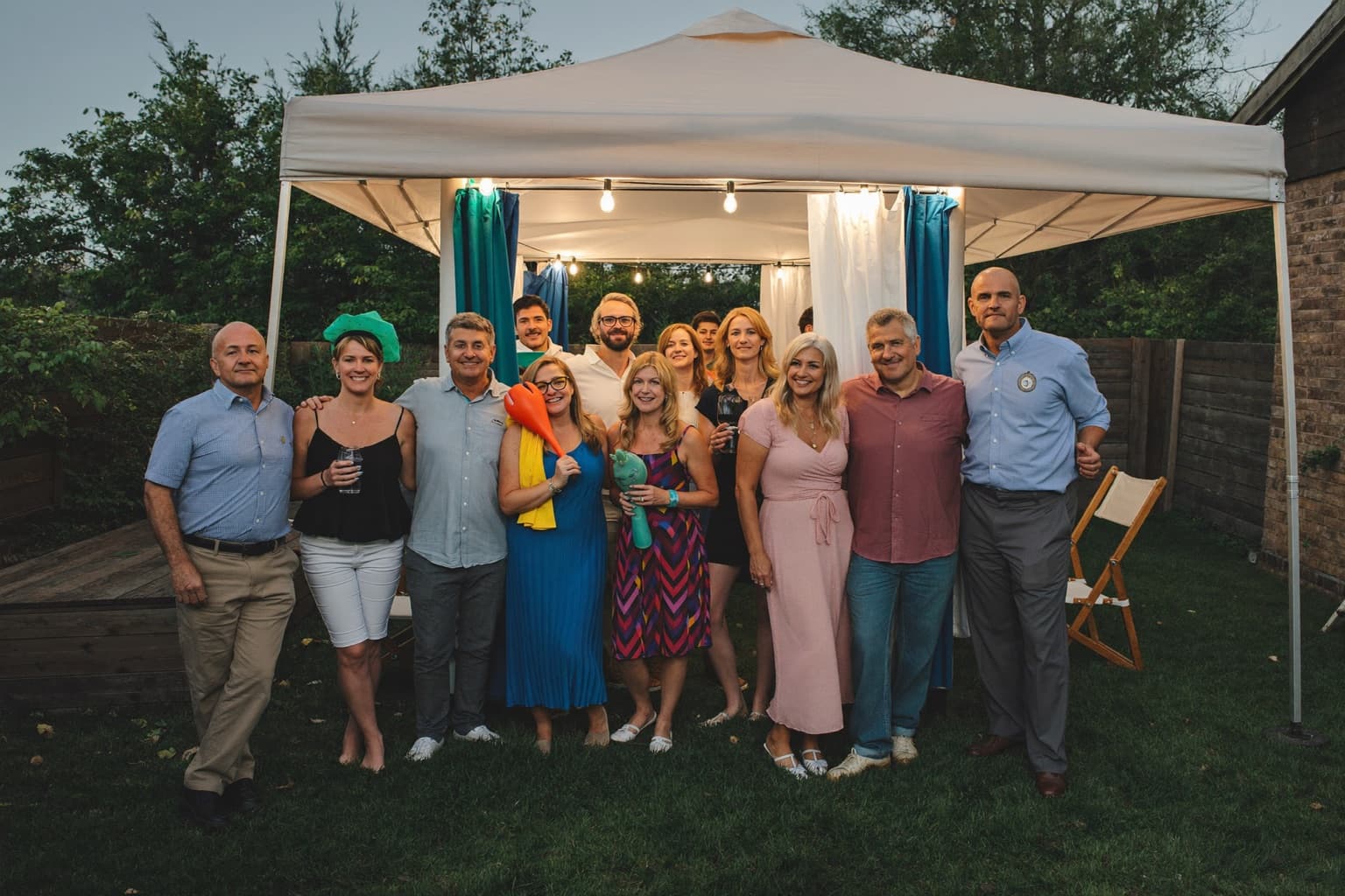 Friends posing with handheld props at an open-air photo booth set up under a backyard party tent