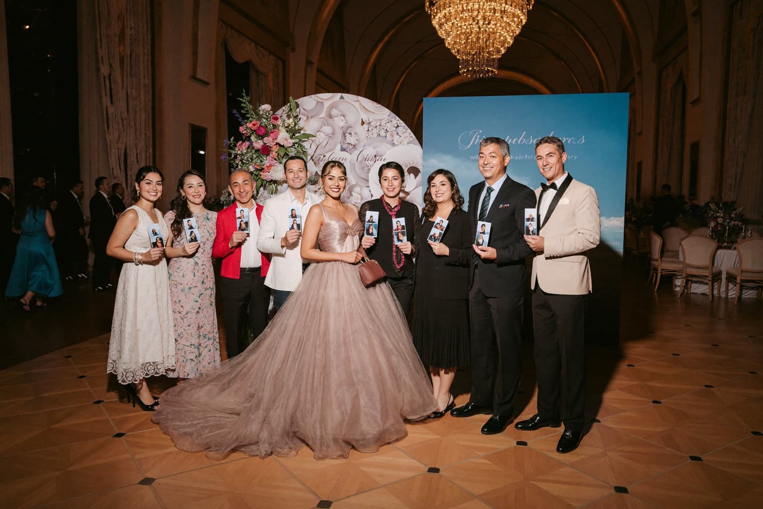 Quinceañera in a formal ball gown posing with friends in front of a gold backdrop at an open-air photo booth