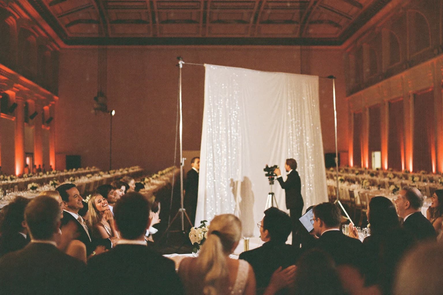 Wedding guests at a white sequin photo booth backdrop in a large uplit banquet-hall ballroom.
