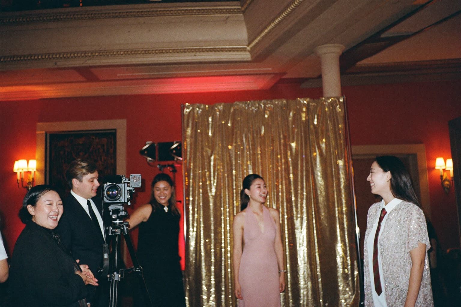 Guests laughing at a gold sequin photo booth backdrop with a camera on a tripod in an ornate theater lobby.