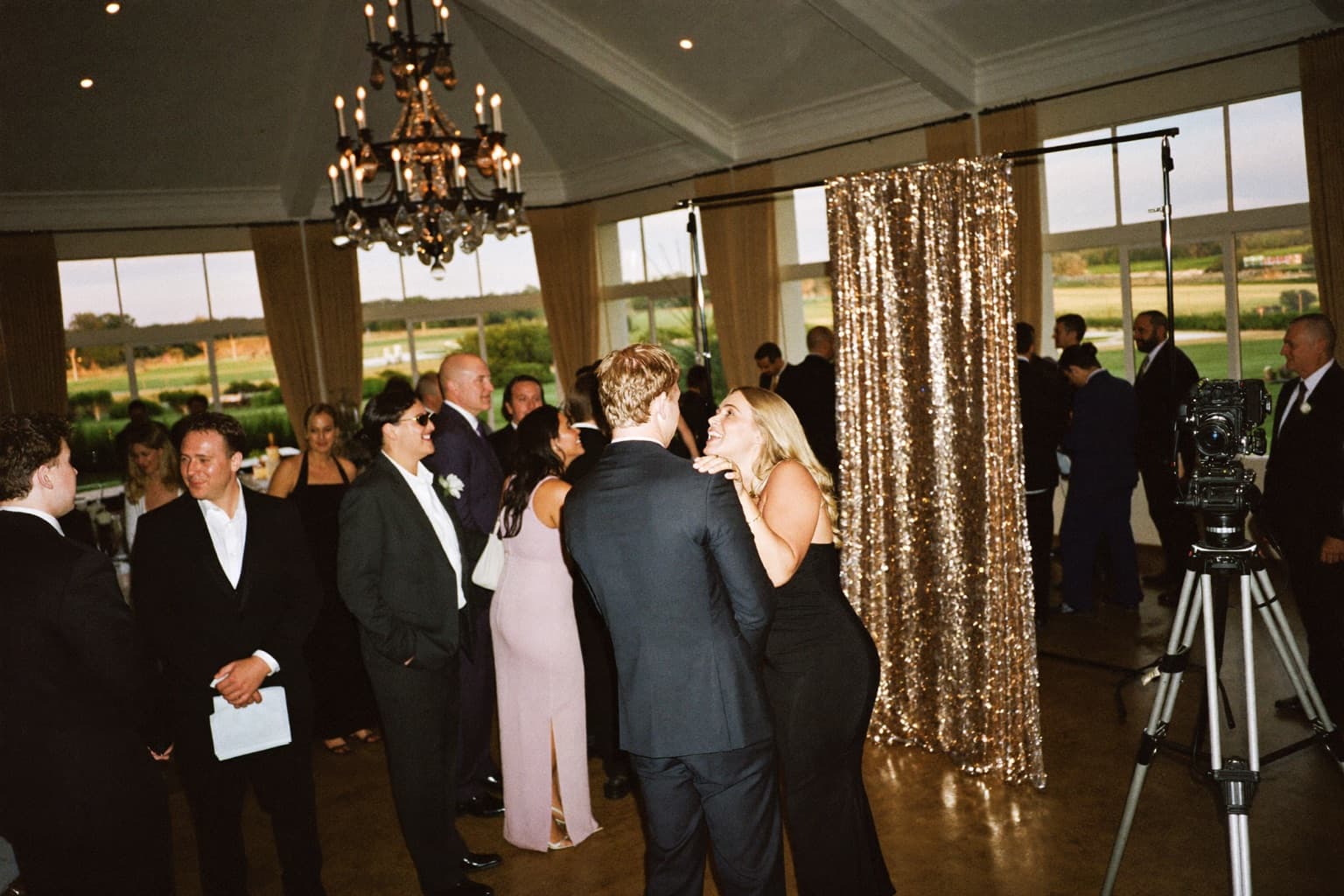 Guests at a champagne sequin photo booth backdrop in a country-club ballroom overlooking a golf course.