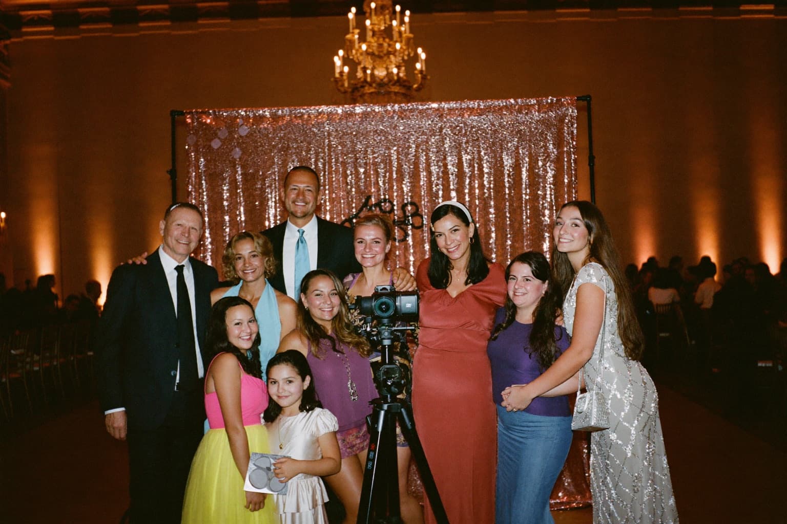 A multigenerational family posing at a rose-gold sequin photo booth backdrop in a grand banquet-hall ballroom.