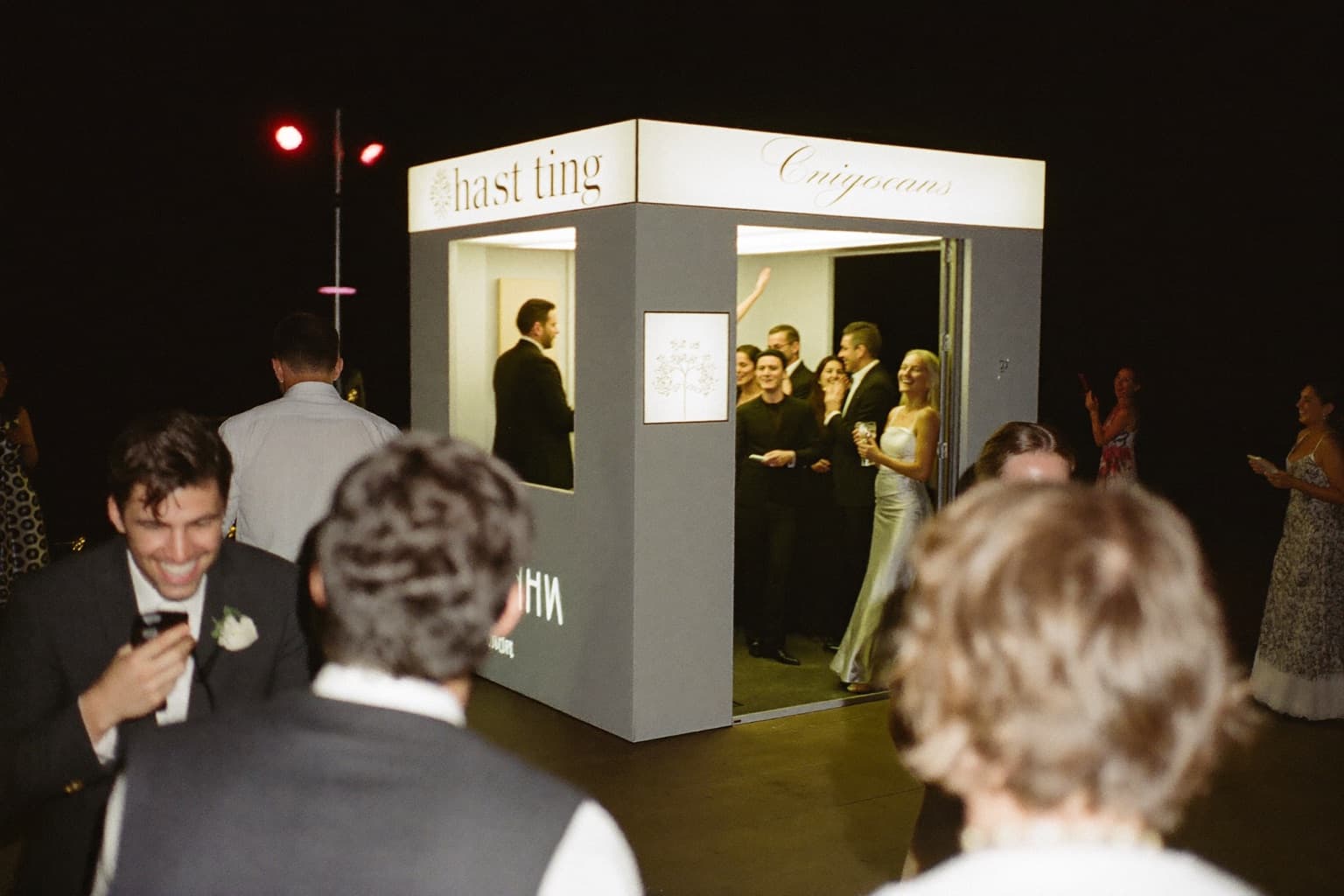 Wedding guests posing together in front of an open-air photo booth backdrop while others dance in the background at a reception