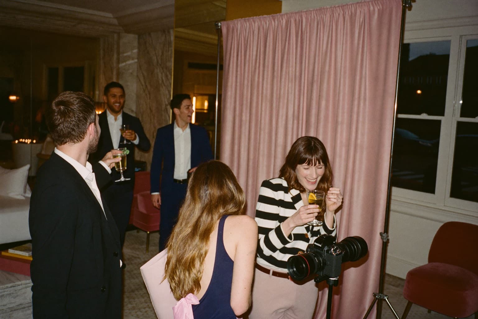 Guests with cocktails at a blush draped photo booth backdrop in a boutique hotel event room at night.