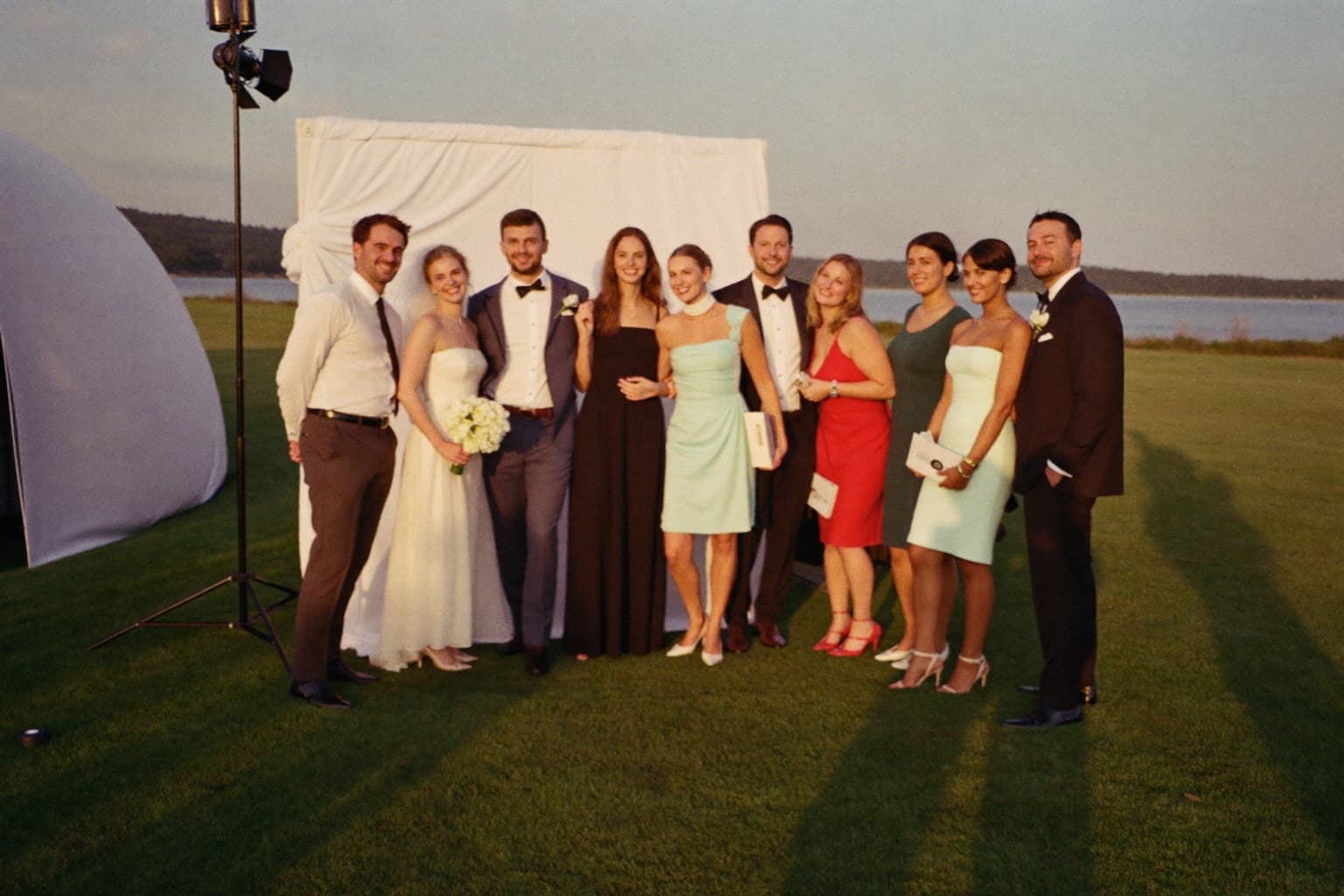 Wedding guests posing at a white photo booth backdrop on a waterfront country-club lawn at golden hour.